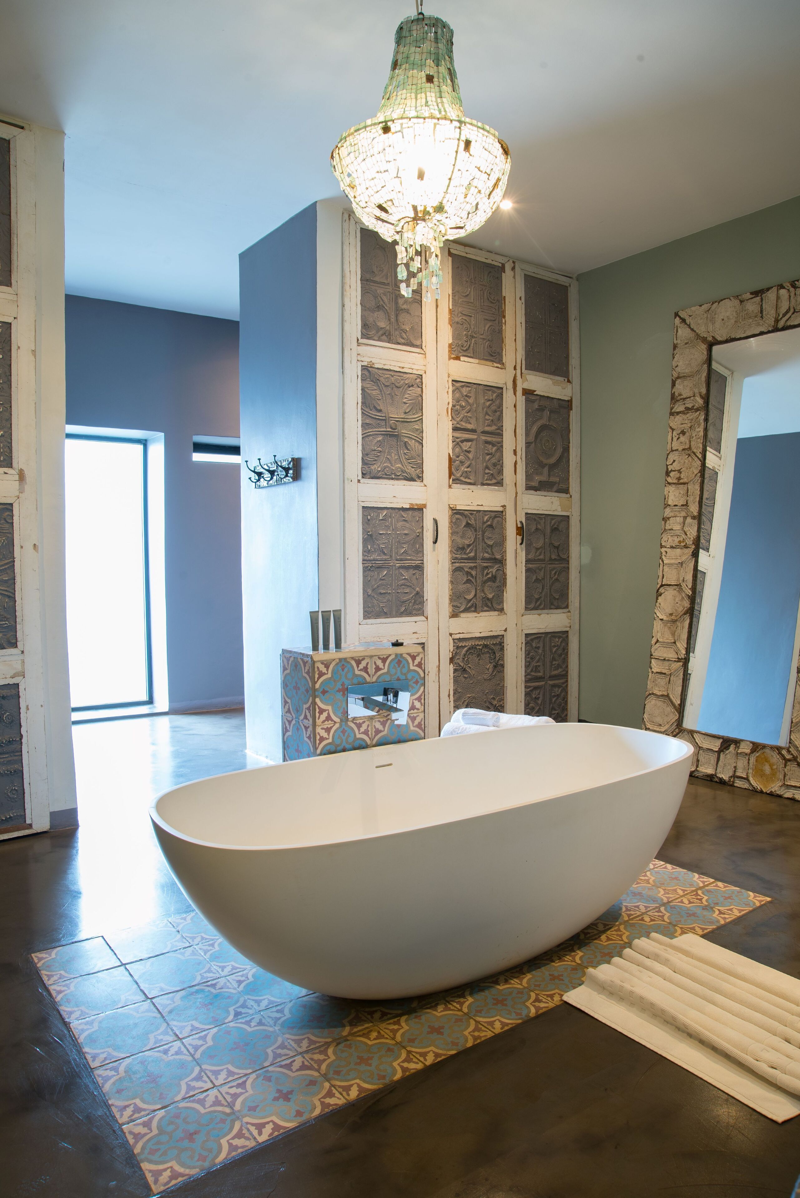 A modern bathroom with a freestanding bathtub, a decorative chandelier, patterned tiles, and a stone accent wall.