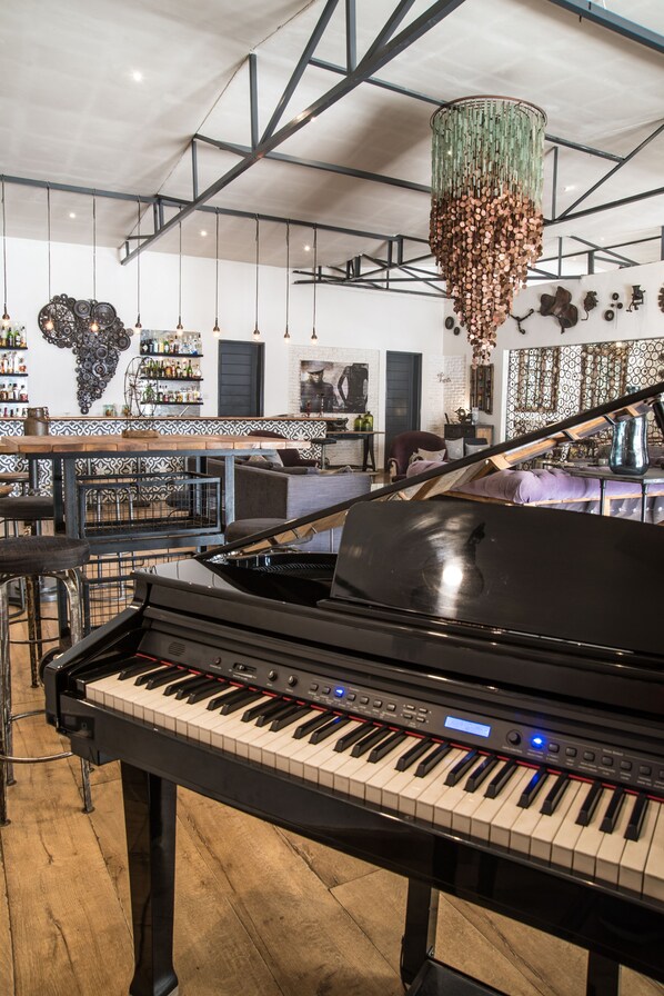 A piano in a spacious room with a high ceiling, a bar area with shelves of bottles, and a unique chandelier. - Latitude 13 Degrees (Lilongwe)