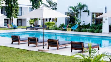Área de piscina com espreguiçadeiras e guarda-sóis, cercada por vegetação e um edifício moderno.