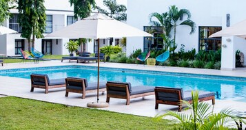 Zona junto a la piscina con sillas de descanso y sombrillas, rodeada de follaje y un edificio moderno.