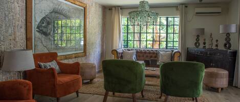 Salón con un cuadro grande de peces, sillas verdes y naranjas, una araña y una ventana con vistas a follaje.