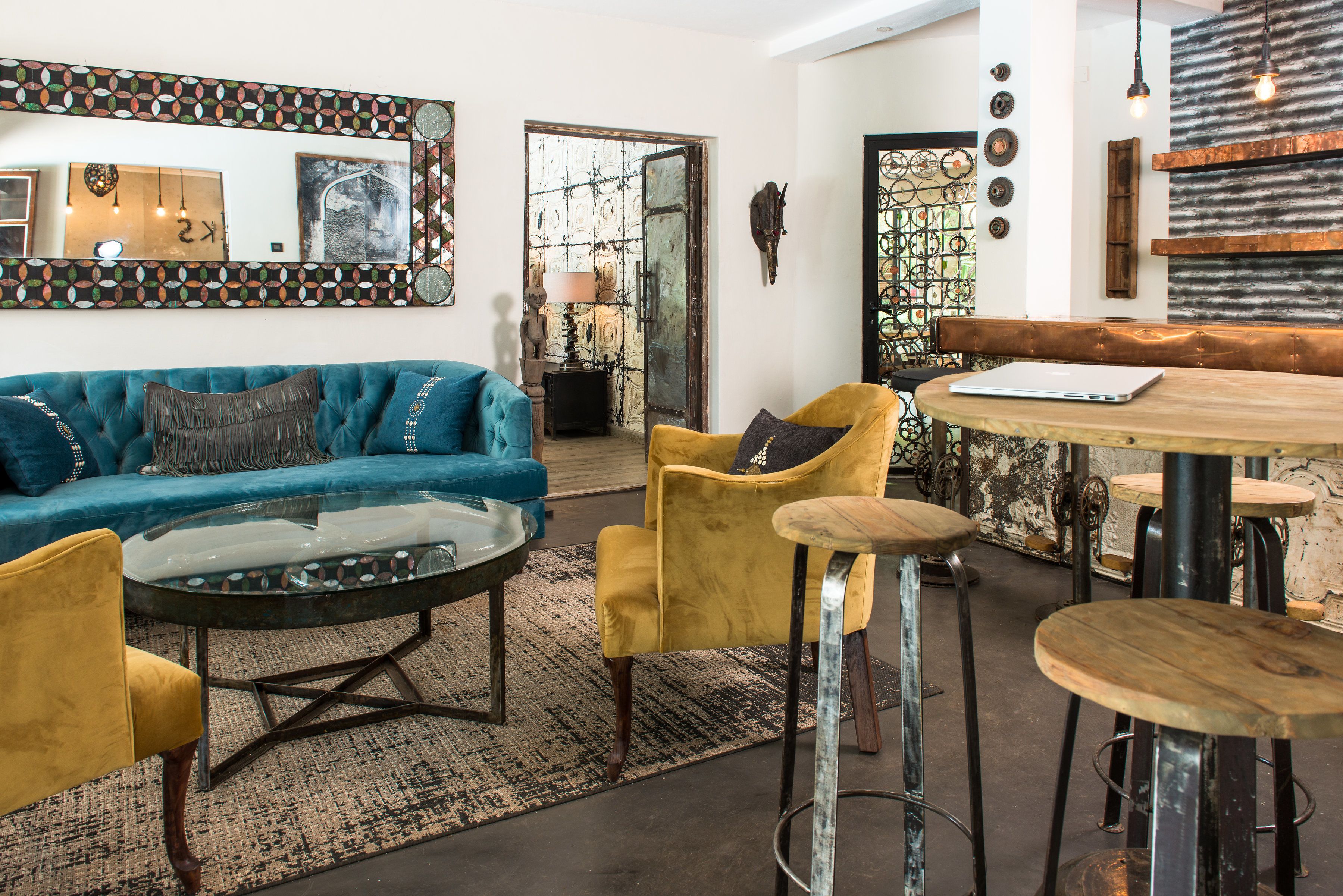 A modern living room with a blue sofa, yellow armchairs, a glass coffee table, and a wooden dining table with bar stools.
