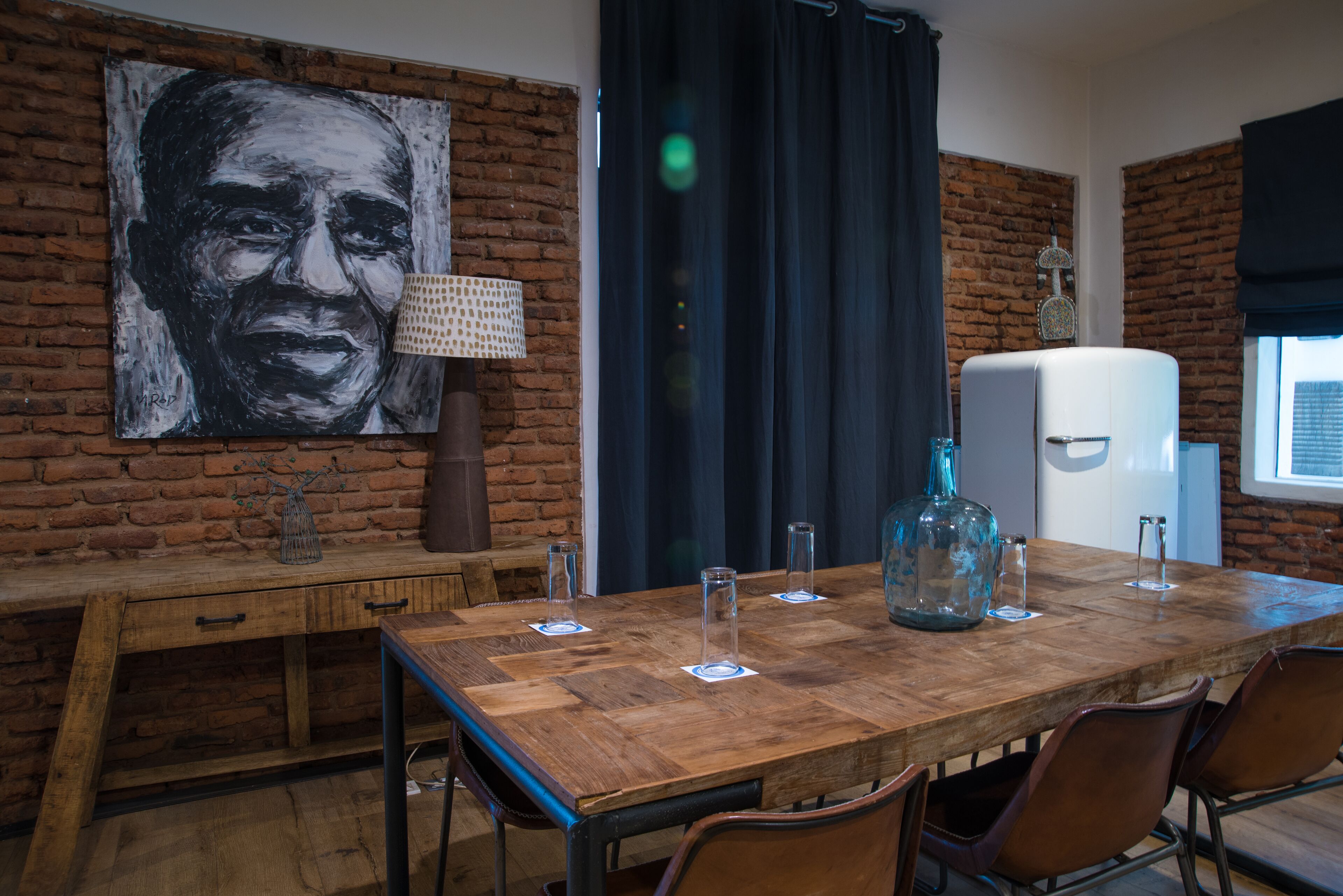 Uma área de refeições com mesa de madeira, cadeiras, um quadro grande na parede, uma luminária e uma geladeira.