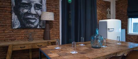 Uma área de refeições com mesa de madeira, cadeiras, um quadro grande na parede, uma luminária e uma geladeira.