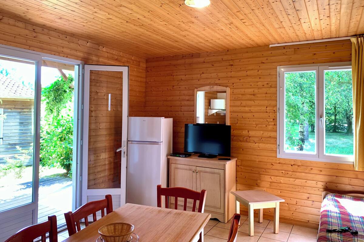 A wooden cabin with a dining area, a refrigerator, a television, and a window with curtains.