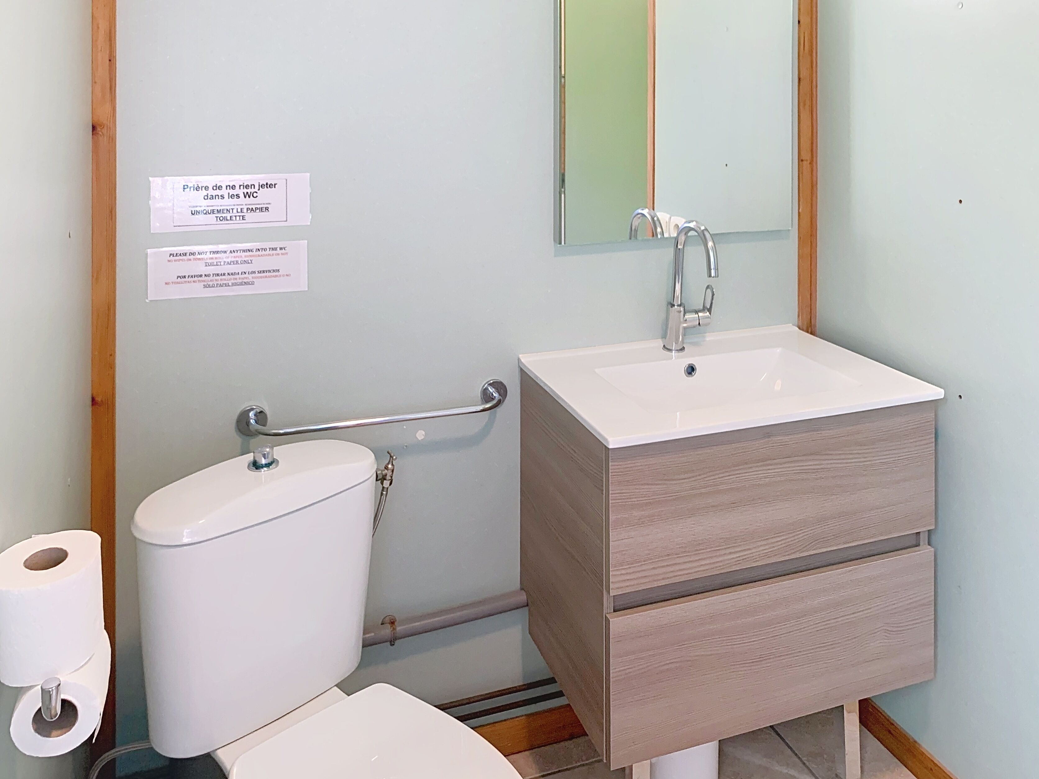 Et badeværelse med toilet, håndvask og spejl. Der er skilte på væggen, og et gelænder er synligt.