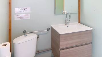 Et badeværelse med toilet, håndvask og spejl. Der er skilte på væggen, og et gelænder er synligt.