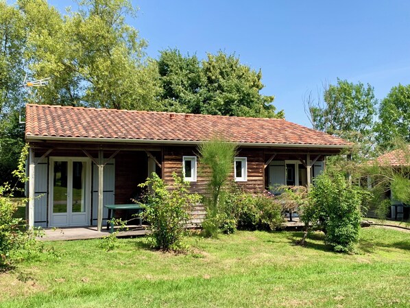 Une maison en bois d’un seul étage, avec un toit en tuiles, une véranda et un petit jardin agrémenté de diverses plantes et arbustes.