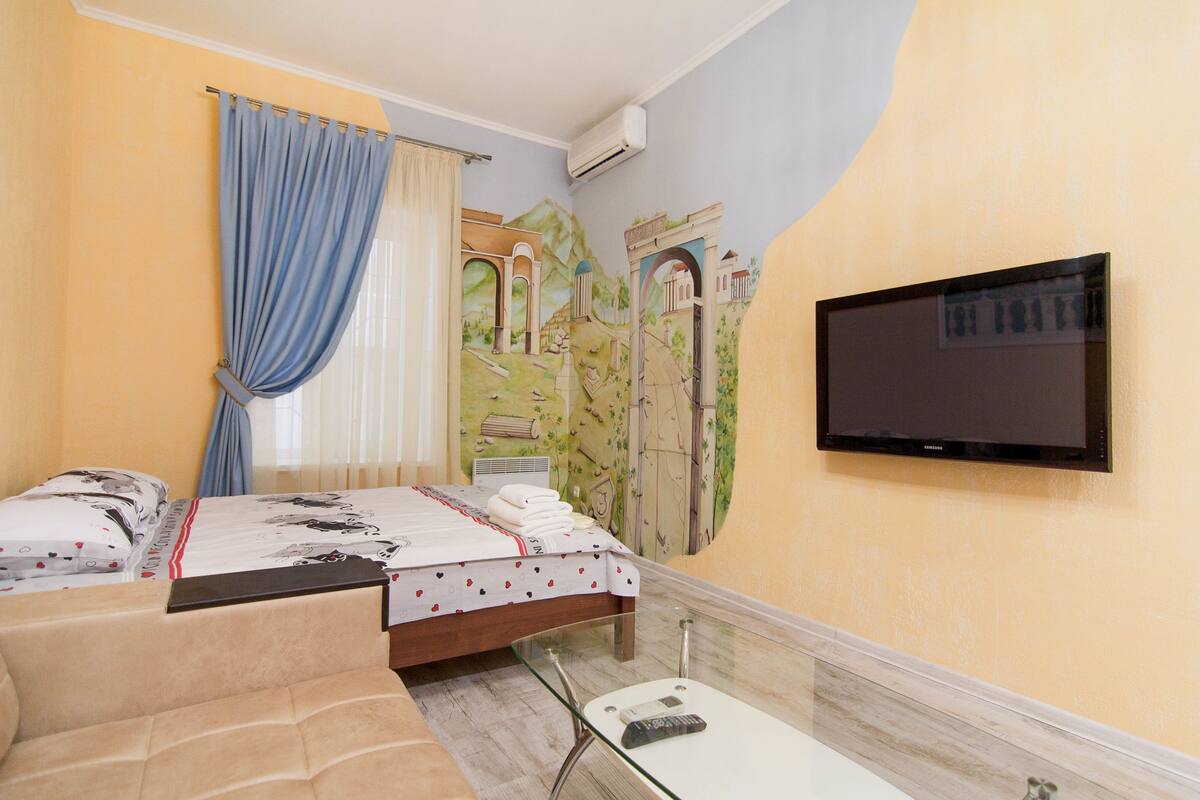A hotel room with a bed, a TV mounted on the wall, and a mural of a landscape.