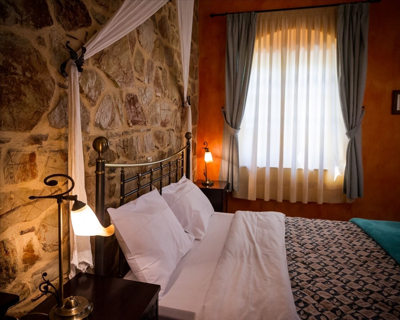 Un lit avec du linge de lit blanc, un mur en pierre, une table de chevet avec une lampe et une fenêtre avec des rideaux.