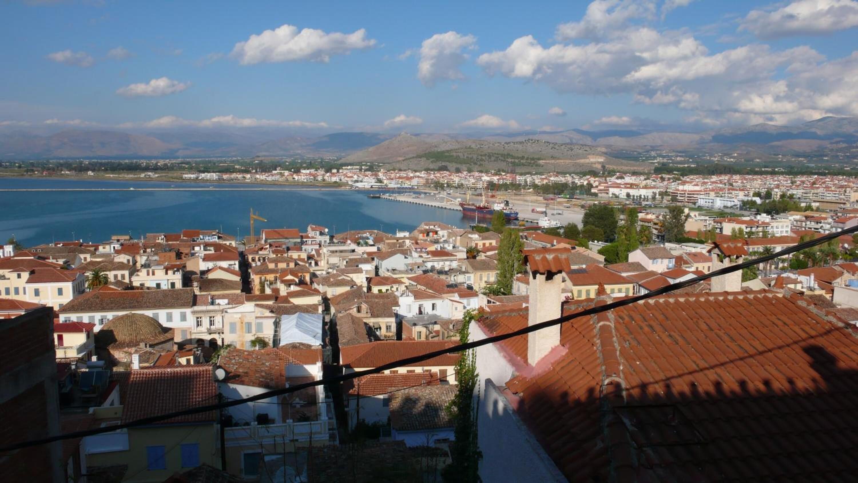 A coastal town with a harbor, red-roofed buildings, and a clear view of the sea and mountains in the distance.