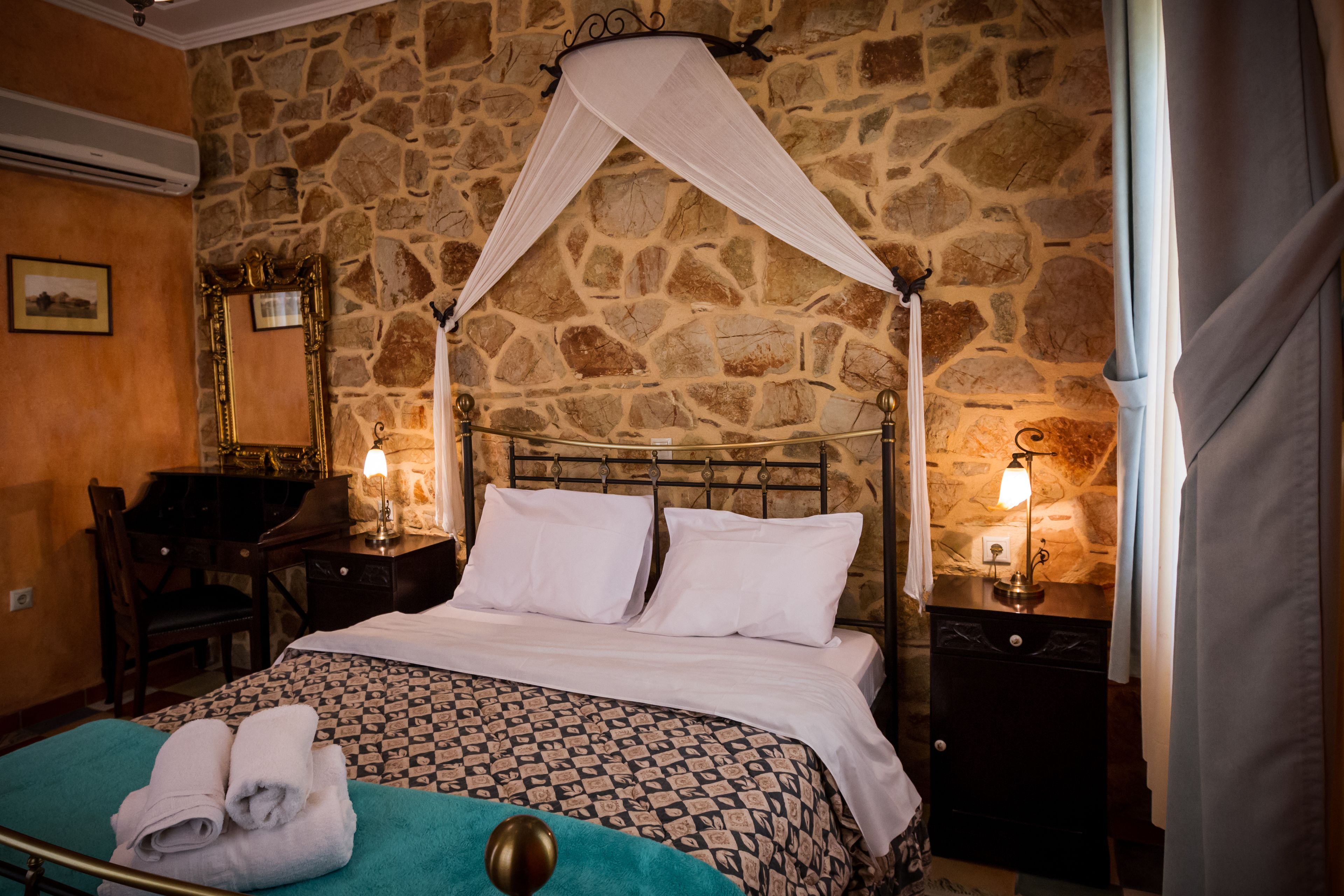 A room with a stone wall, a bed with a canopy, a desk with a chair, and two bedside lamps.