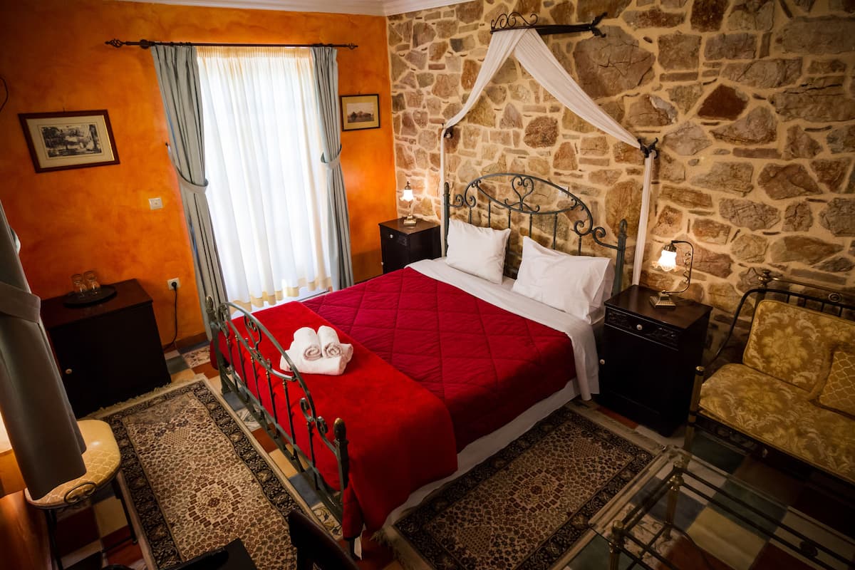 A bedroom with a stone wall, a bed with a red quilt, a nightstand with a lamp, and a chair.