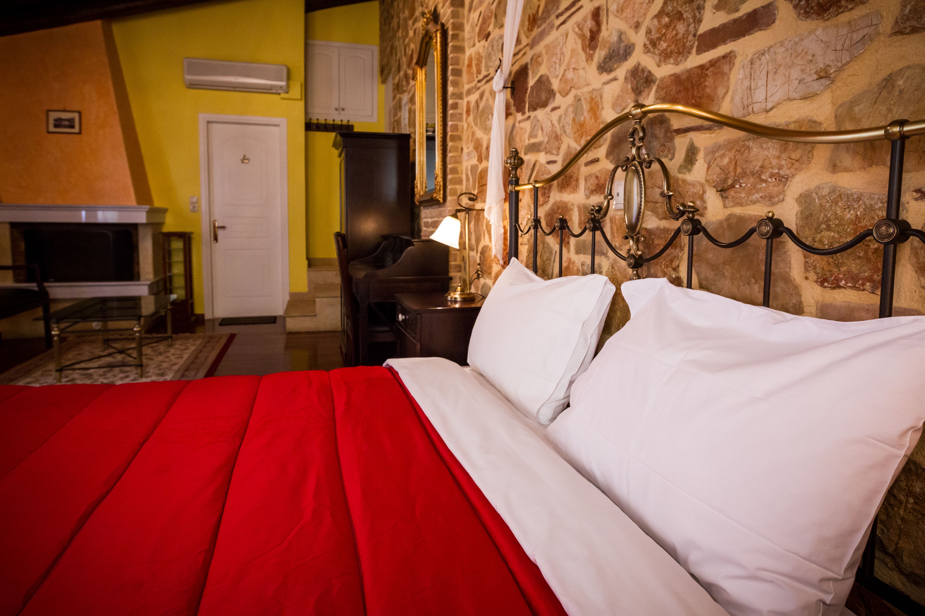 A bed with a red bedspread and white pillows, a stone wall, a wooden desk, a lamp, and a white door.