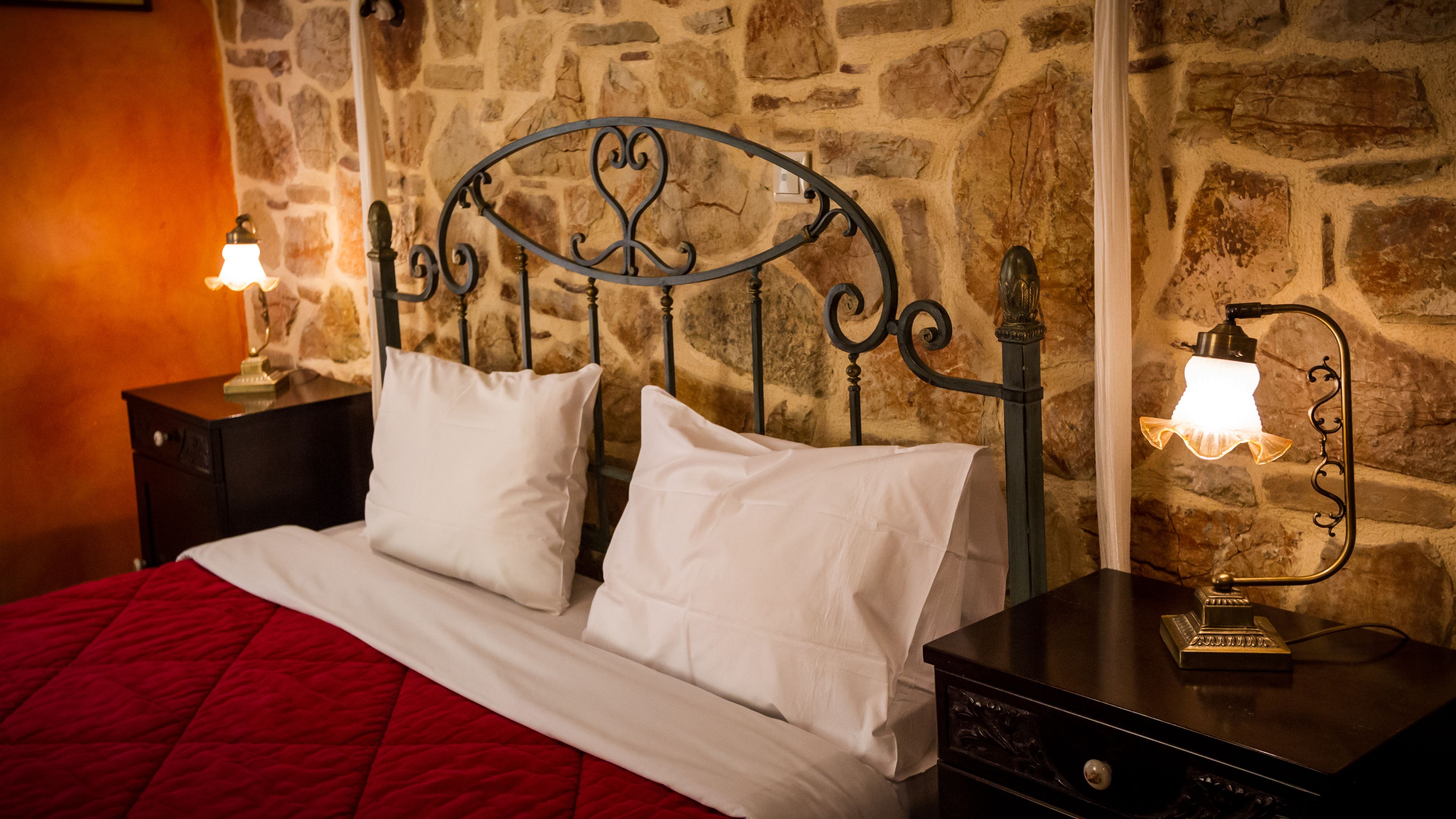 Una cama con sábanas blancas, una colcha roja y un cabecero metálico frente a una pared de piedra. Hay dos mesitas de noche con lámparas.