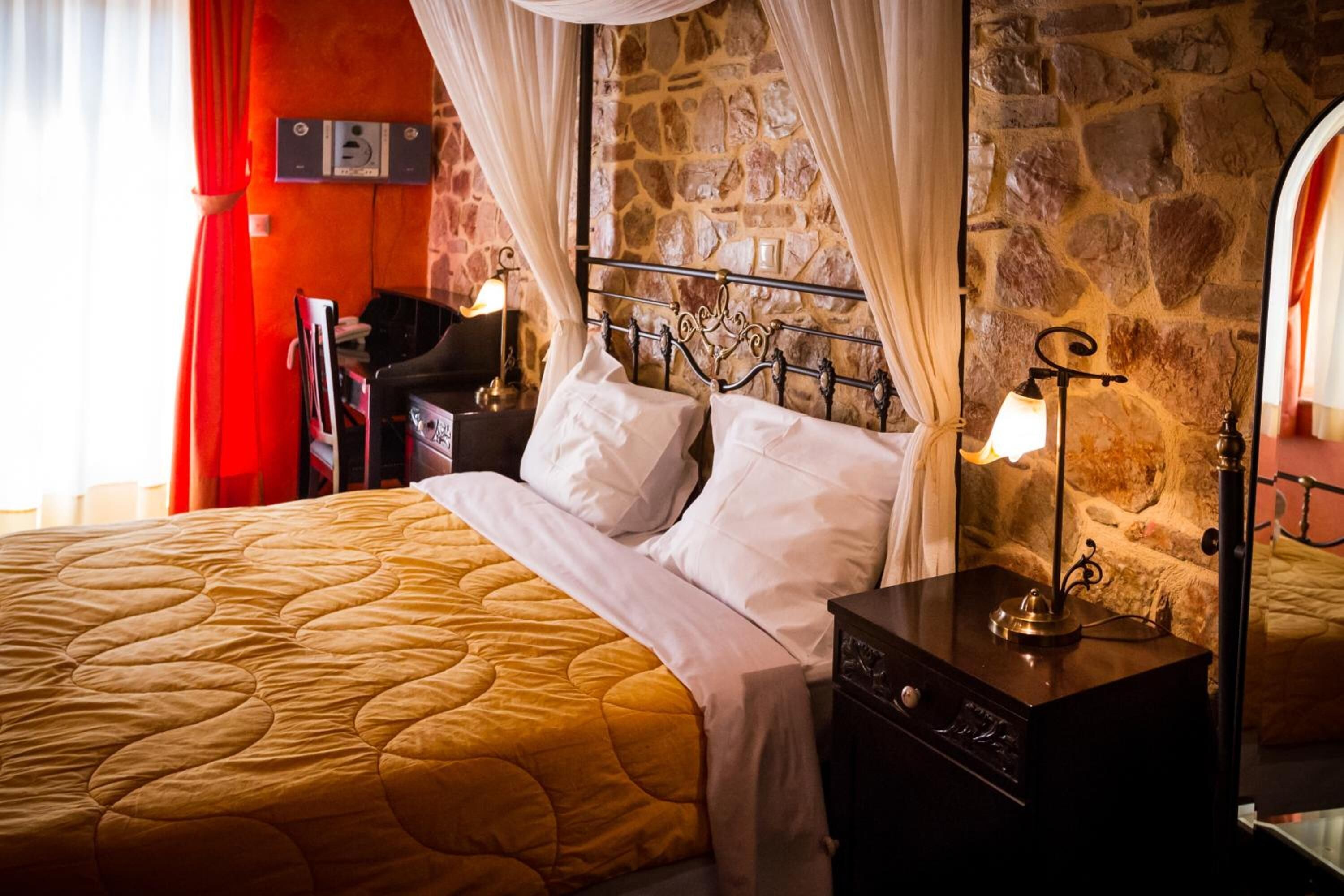 A bed with a yellow quilt, white pillows, and a metal headboard. There is a nightstand with a lamp and a small table with a lamp. The room has a stone wall and a red curtain.