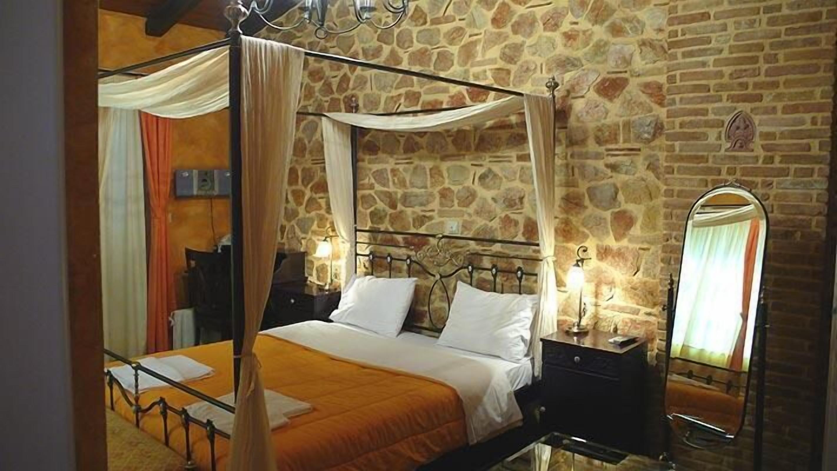 A four-poster bed with a stone wall backdrop, a bedside table with a lamp, and a full-length mirror.