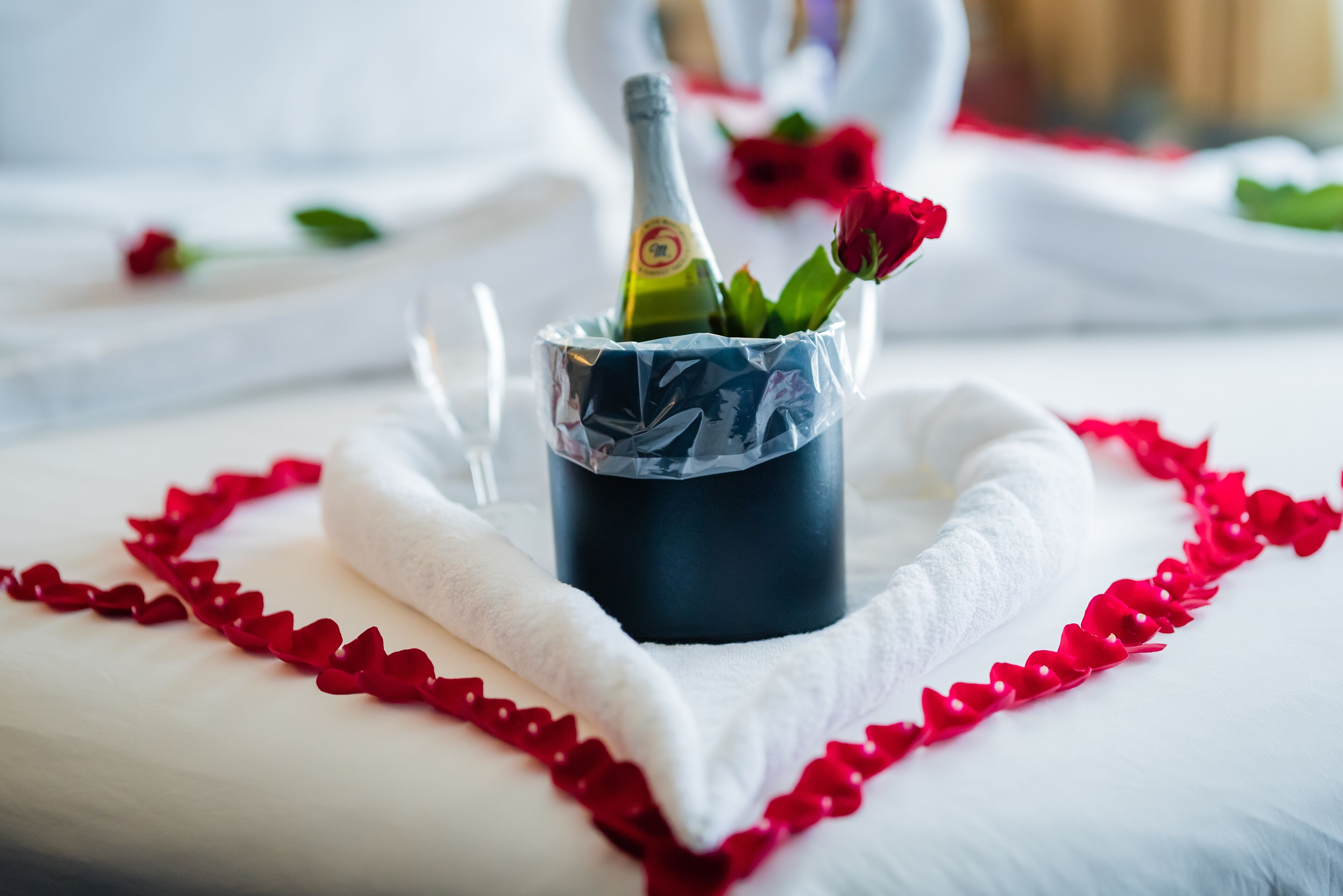Un arrangement de roses rouges en forme de cœur sur une surface blanche, une bouteille de champagne dans un présentoir décoratif et une serviette blanche.