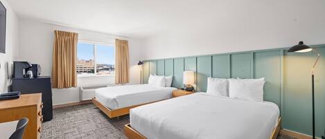 A hotel room with two beds, a desk, a chair, a lamp, and a view of the city.
