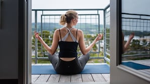 Eine Person sitzt im Schneidersitz auf einer Yogamatte in einem Raum mit Blick auf Grünflächen und Hügel durch ein Metallgitter.