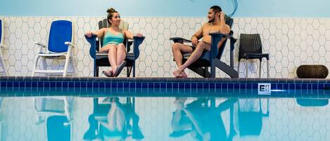 Dos personas sentadas junto a una piscina, con un muro de azulejos y un suelo azul a la vista.