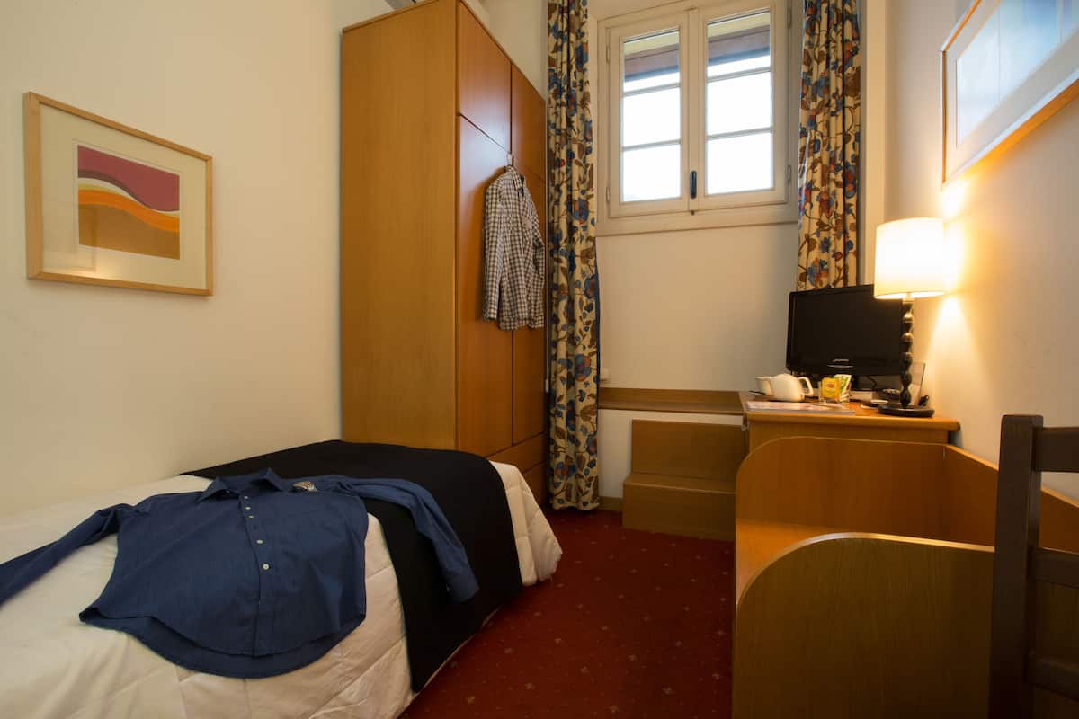 A hotel room with a bed, a desk with a lamp, a wardrobe, and a window with curtains.