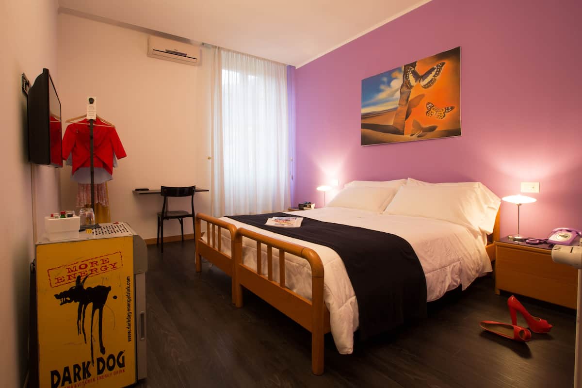 A hotel room with a bed, a chair, a desk with a lamp, a coat rack, and a poster on the wall.