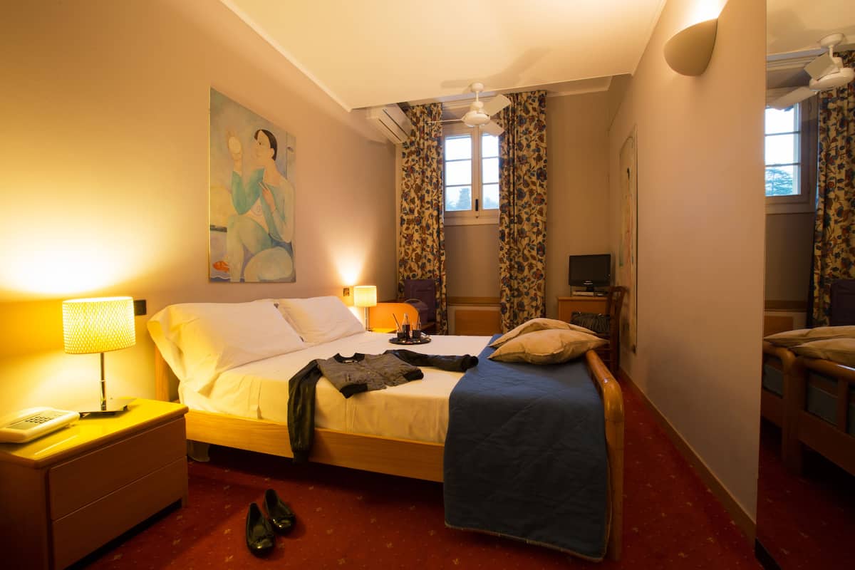 A hotel room with a bed, a nightstand with a lamp, a painting on the wall, and a window with curtains.