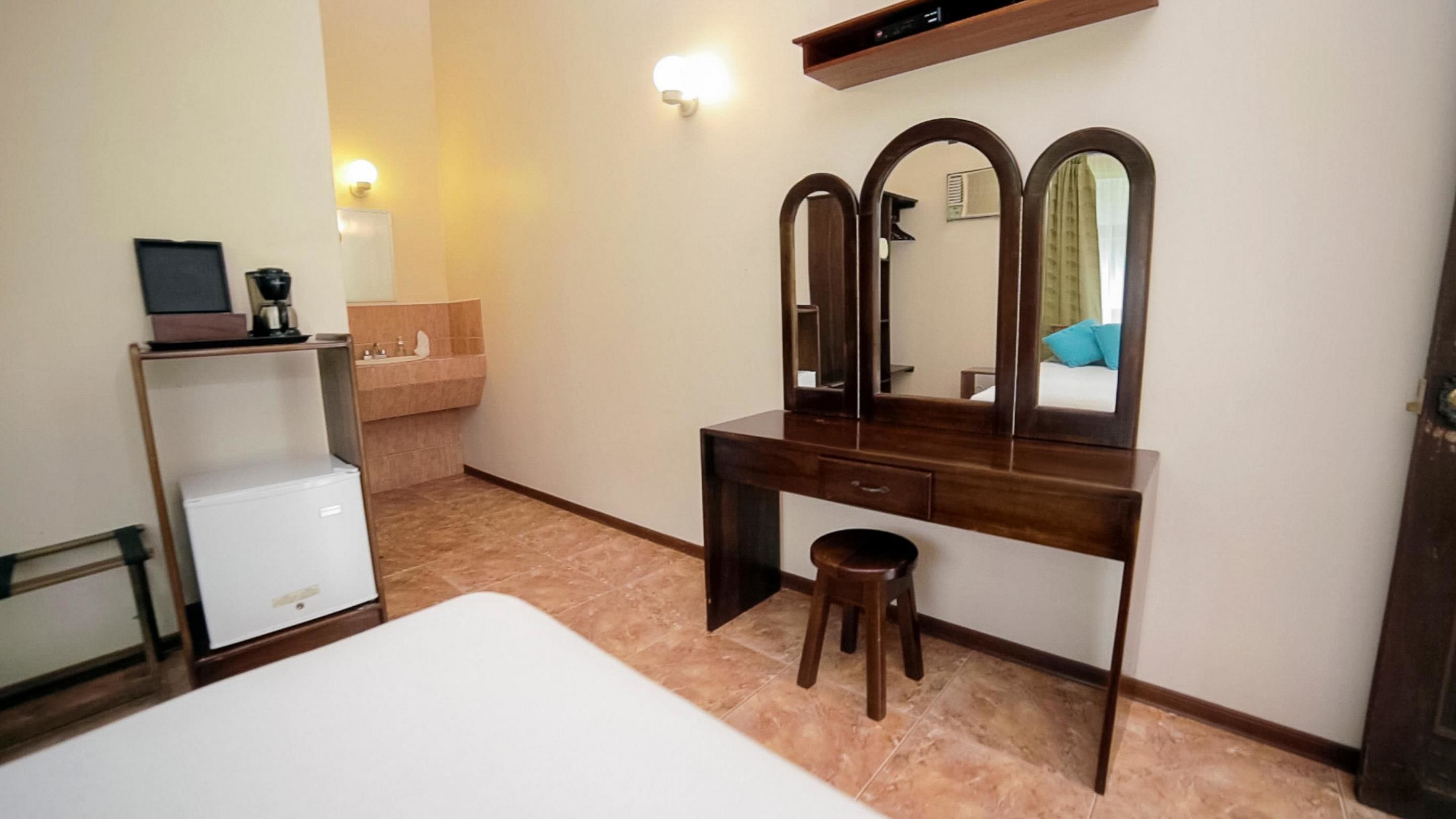 Une chambre d’hôtel comprenant un lit, une commode en bois avec un miroir, un petit tabouret, une armoire blanche et une étagère fixée au mur.