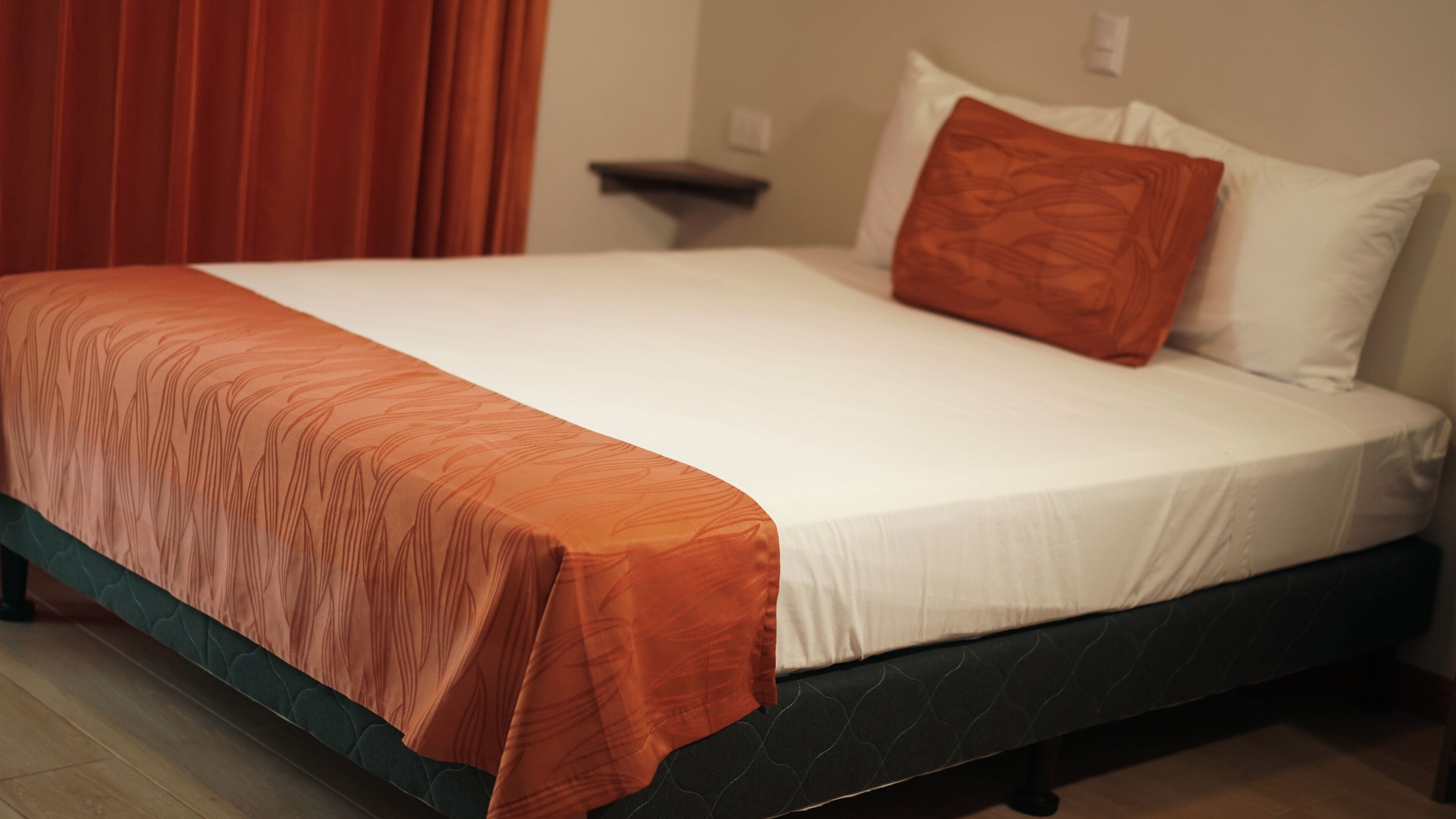 A bed with a white mattress and an orange bedspread, a dark headboard, and a red curtain in the background.