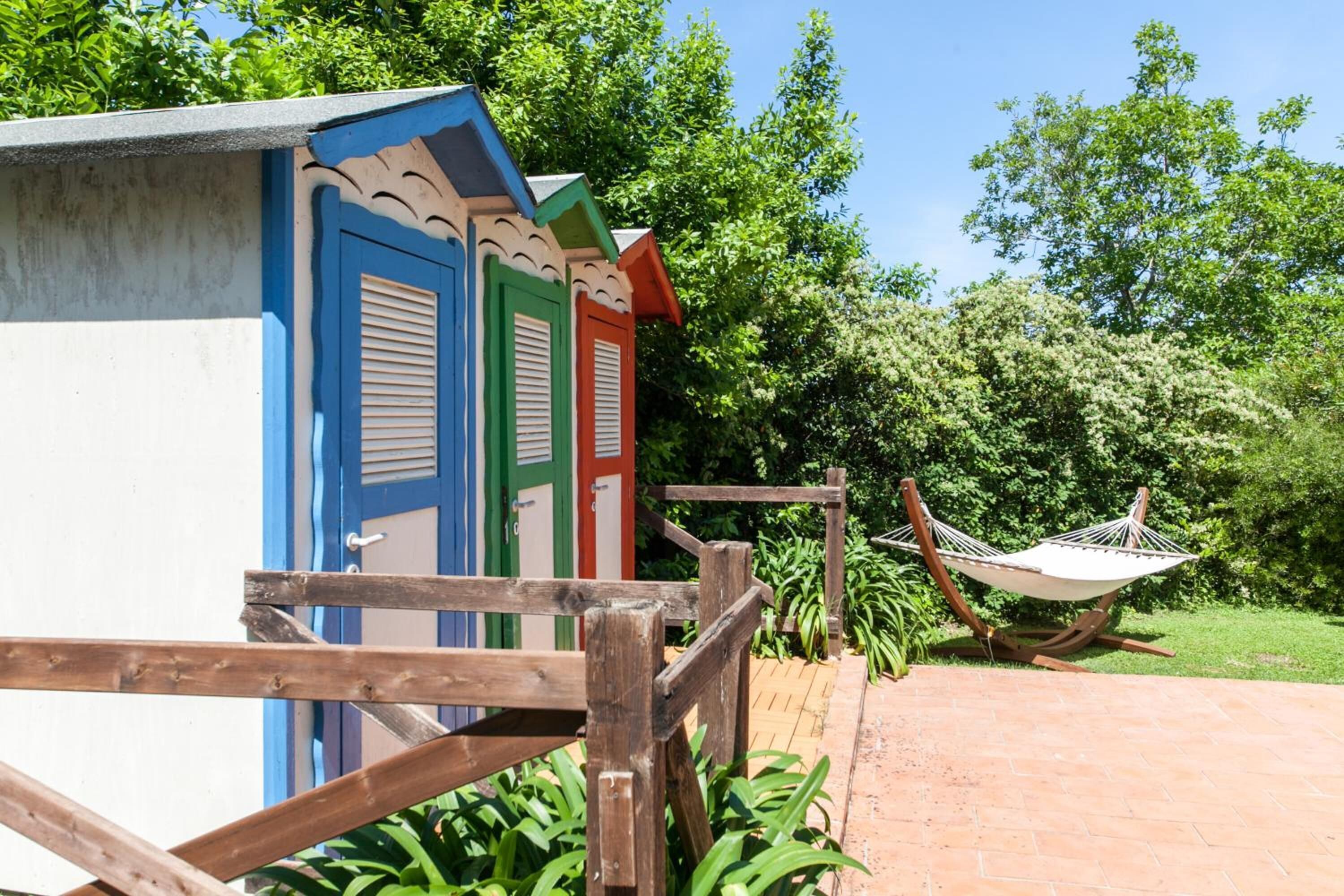 A wooden fence leading to colorful, single-story buildings with shutters, a hammock, and lush greenery in the background.