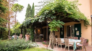 A covered outdoor seating area with wicker chairs and tables, surrounded by greenery and a brick pathway. - Locanda Antiche Macine (Santarcangelo di Romagna)