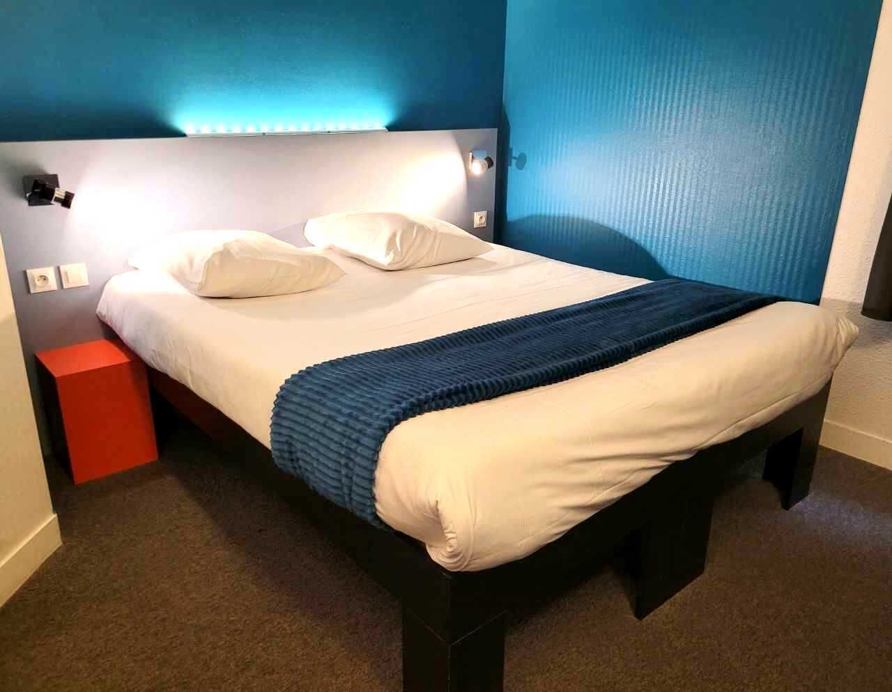Un lit avec une literie blanche et une tête de lit bleue, une table de chevet rouge et un mur bleu.