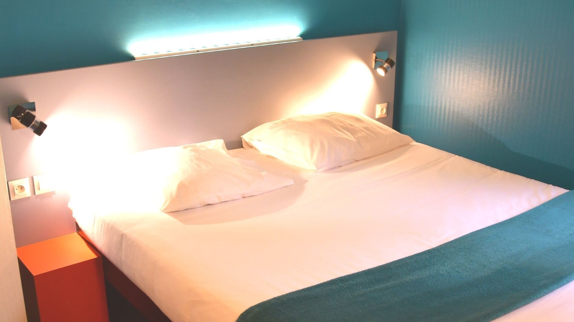 Un lit avec une literie et des oreillers blancs, un plaid turquoise, une table de chevet rouge et une lampe fixée au mur.