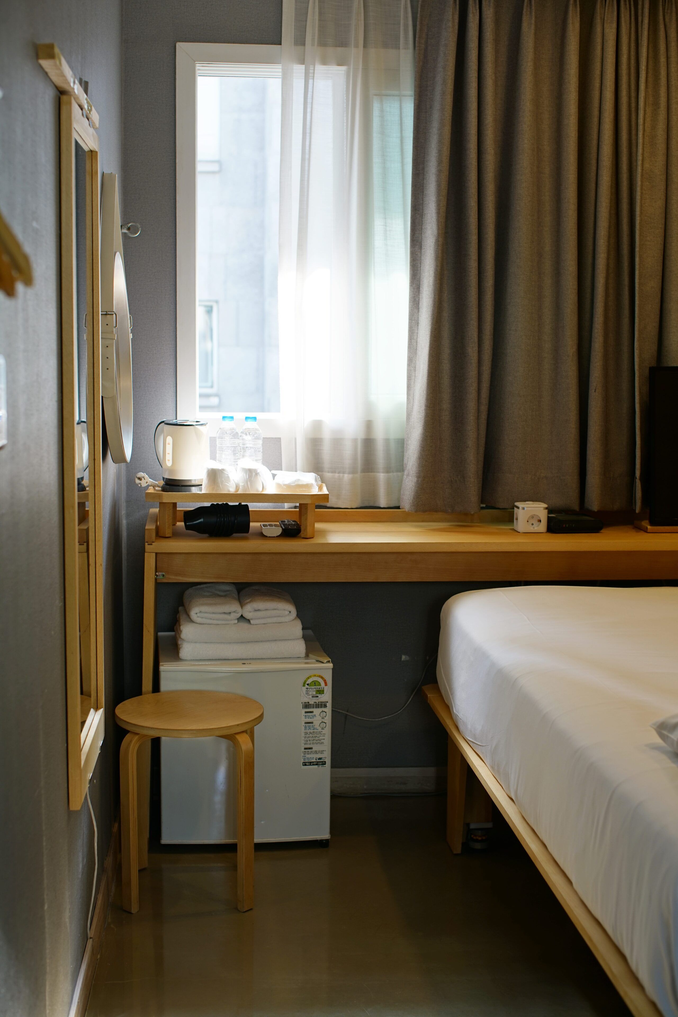 Une chambre d’hôtel comprenant un lit, un bureau avec un ordinateur, un petit réfrigérateur, une fenêtre avec des rideaux et un tabouret en bois.