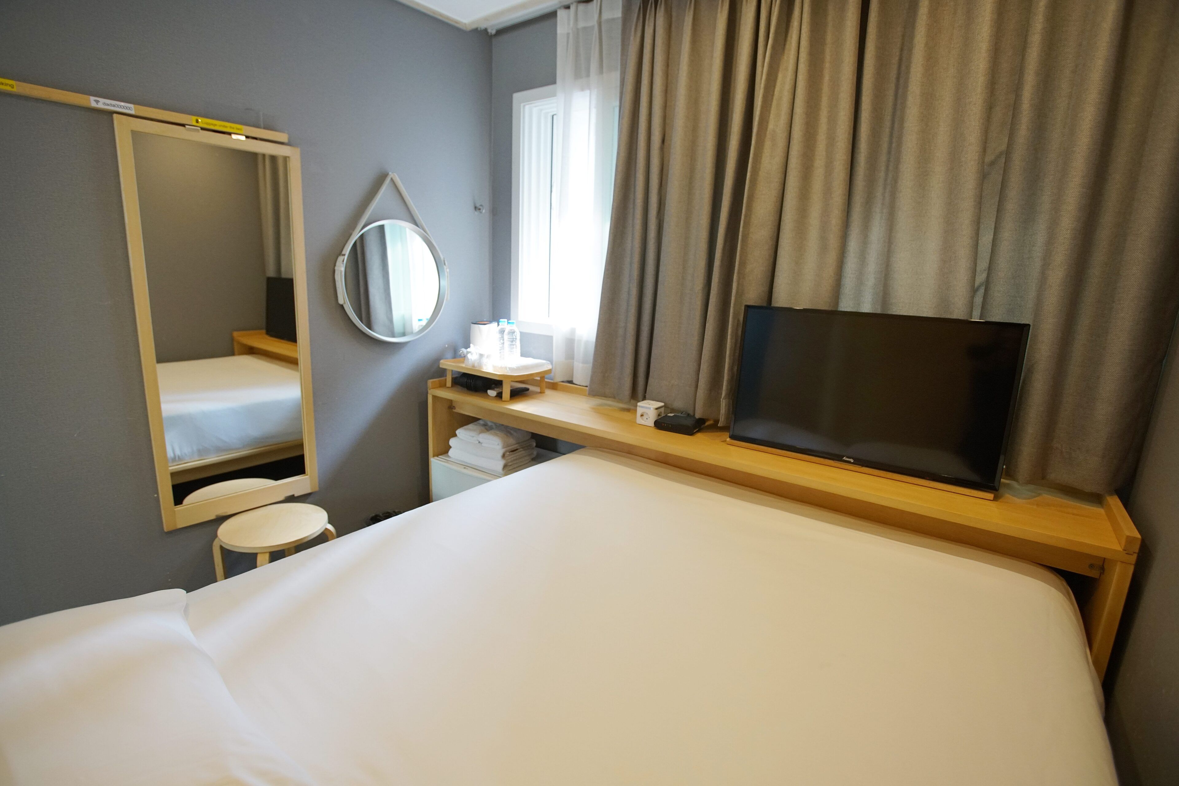 A hotel room with a bed, a TV, a mirror, a small table, and a window with curtains.