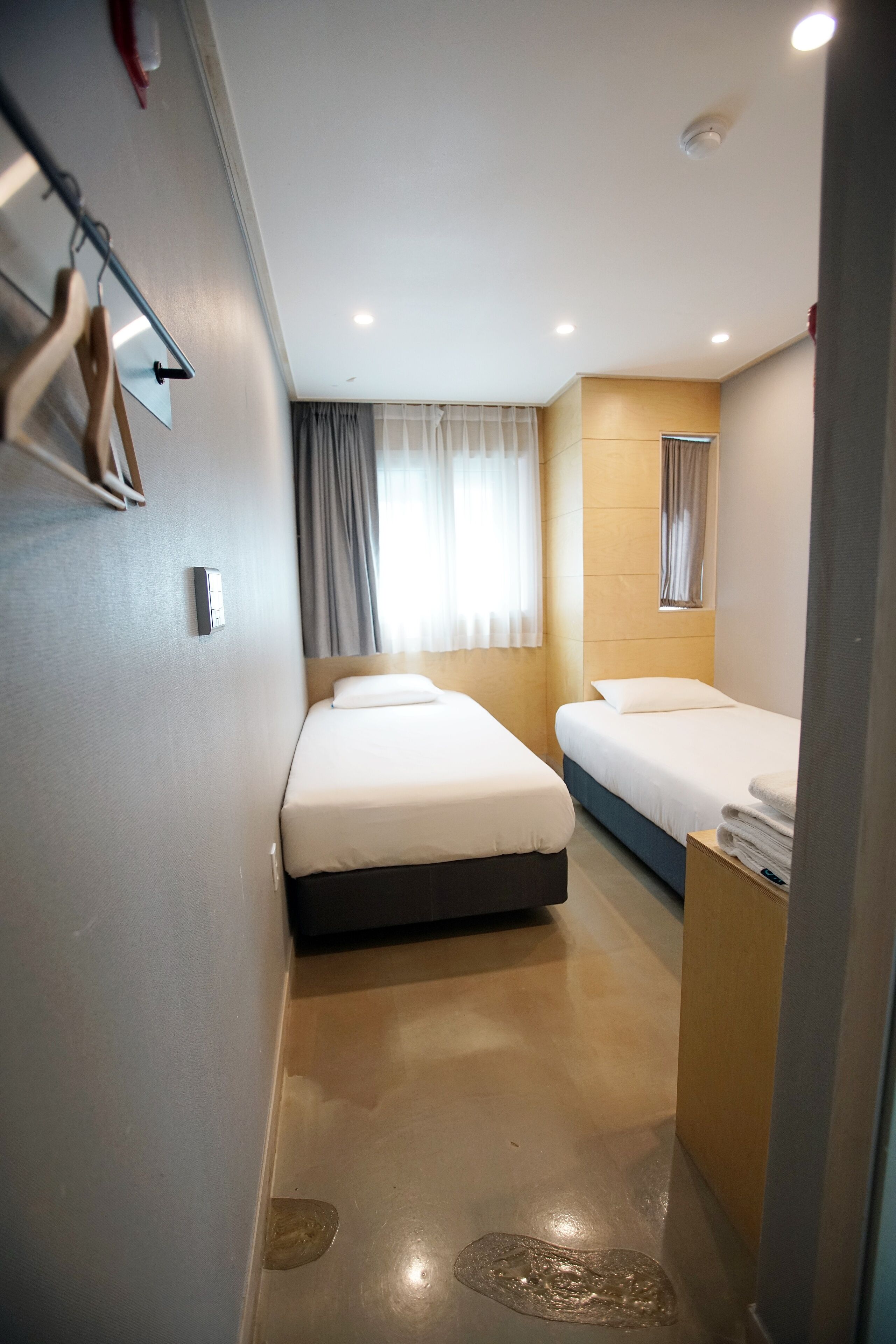 Habitació d'hotel amb dos llits, un capçal de llit de fusta, una finestra petita amb cortines, un mirall i una taula de nit de fusta amb tovalloles.
