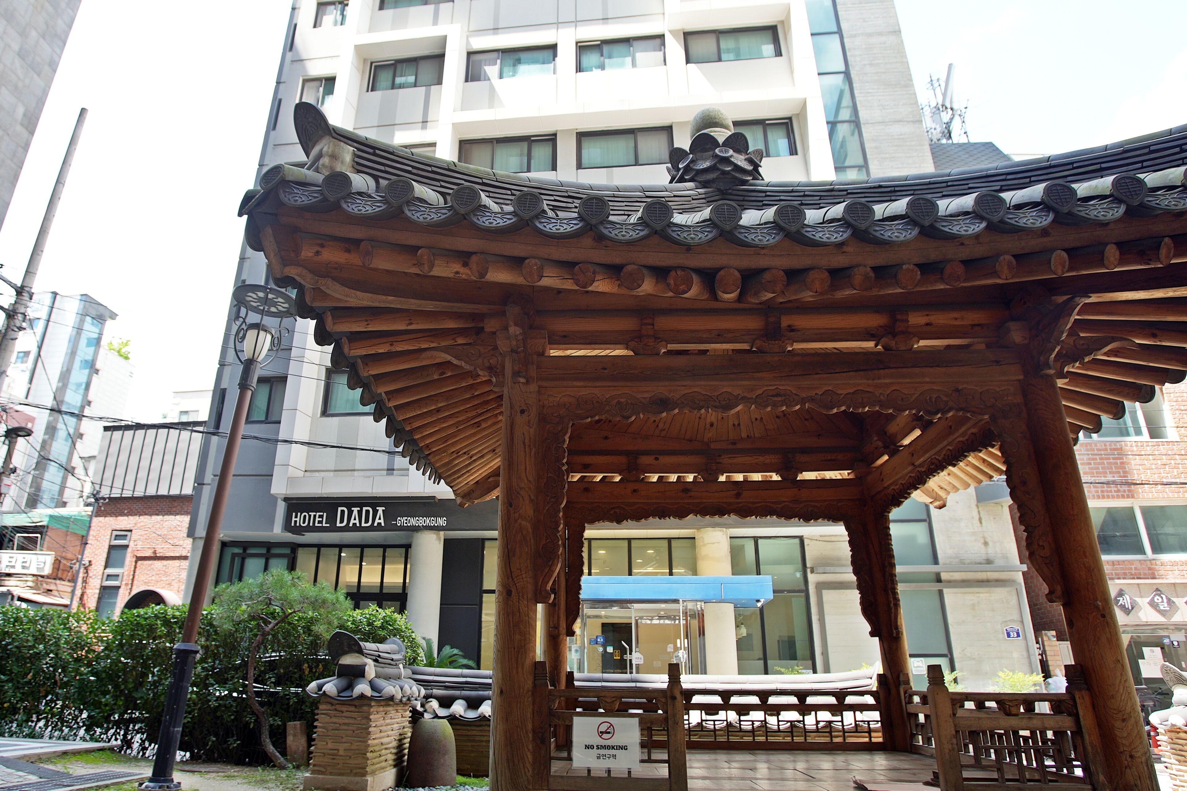 A traditional wooden pavilion with intricate roof design, located in an urban setting with modern buildings in the background.