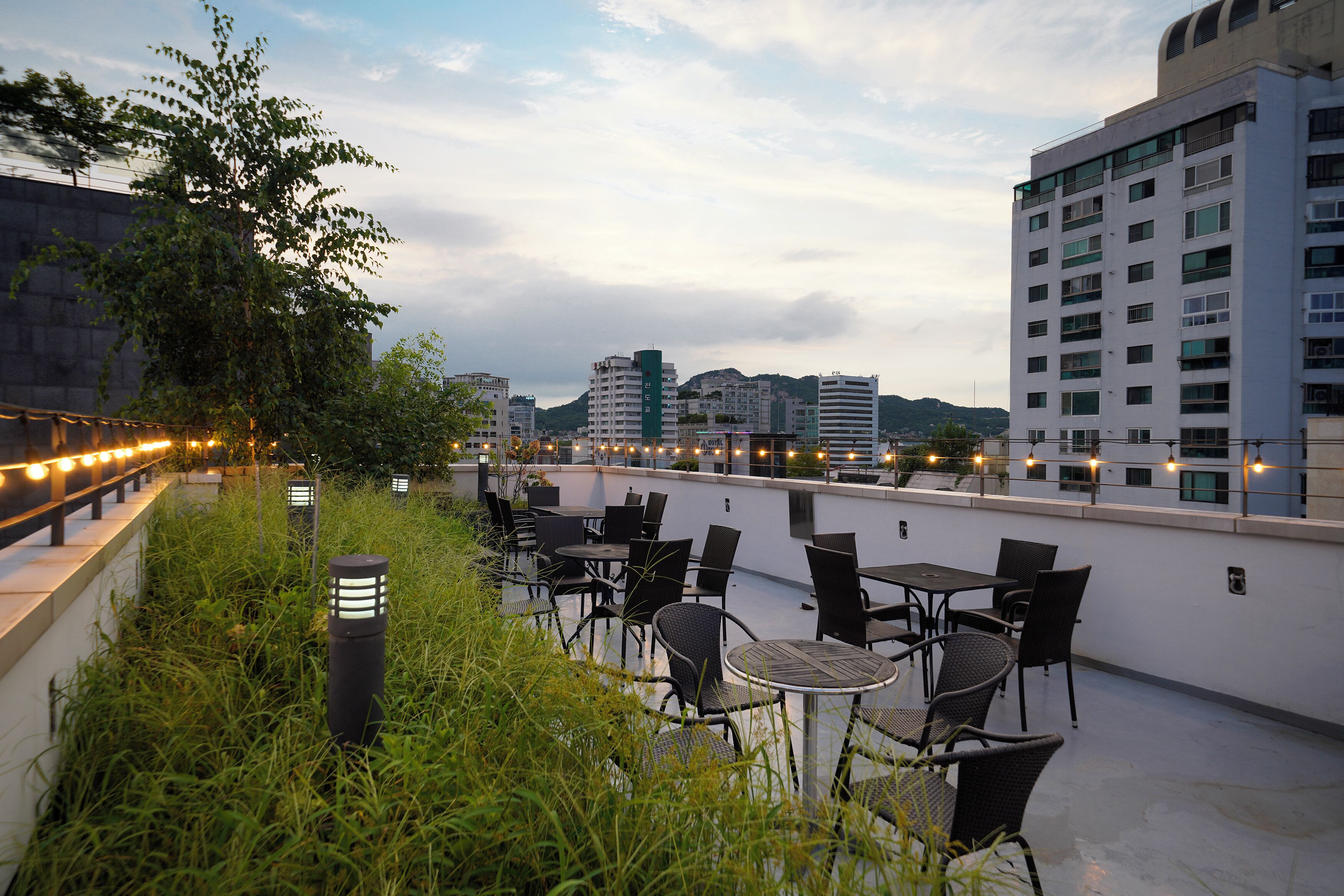 Une terrasse extérieure avec des tables et des chaises, entourée de verdure et avec des bâtiments de la ville en arrière-plan.