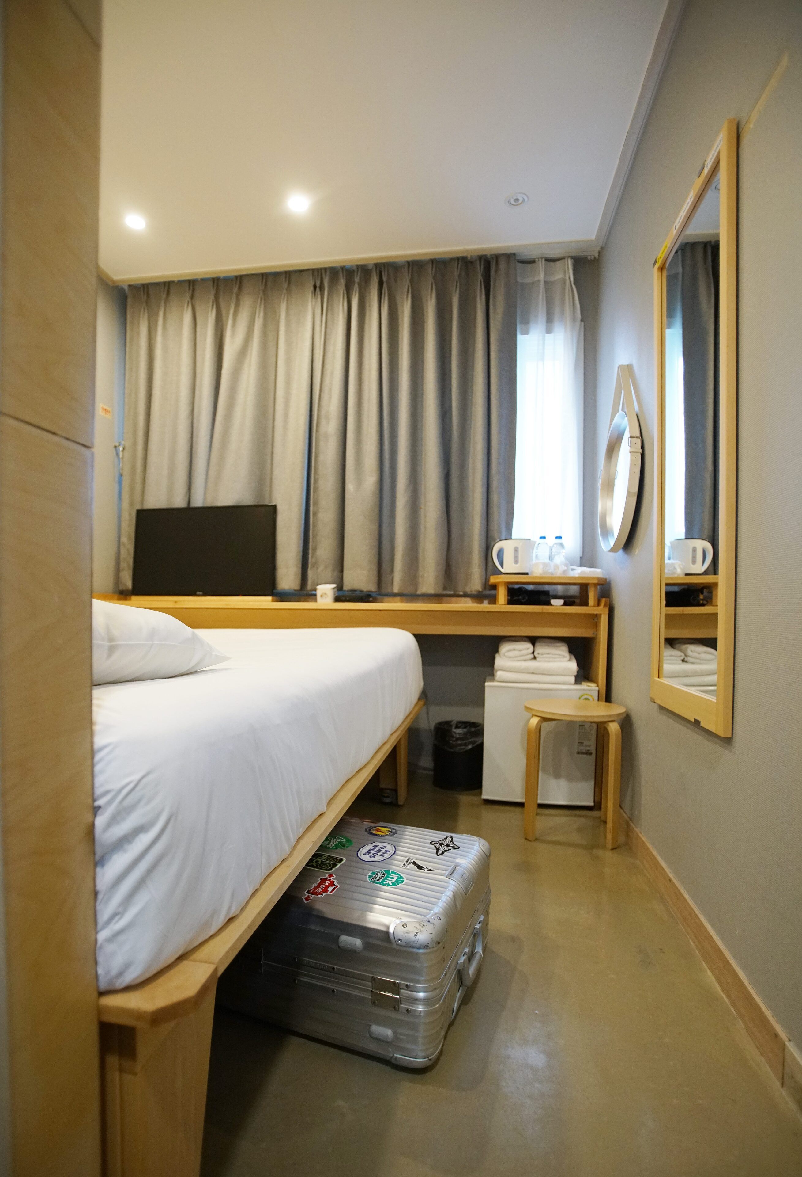 Habitació d'hotel petita amb un llit individual, una taula de treball amb un ordinador, una taula petita, un mirall i una maleta.