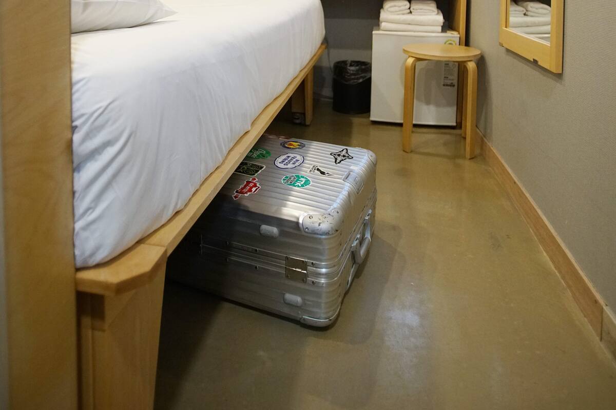 A compact hotel room with a single bed, a desk with a computer, a small table, a mirror, and a suitcase.