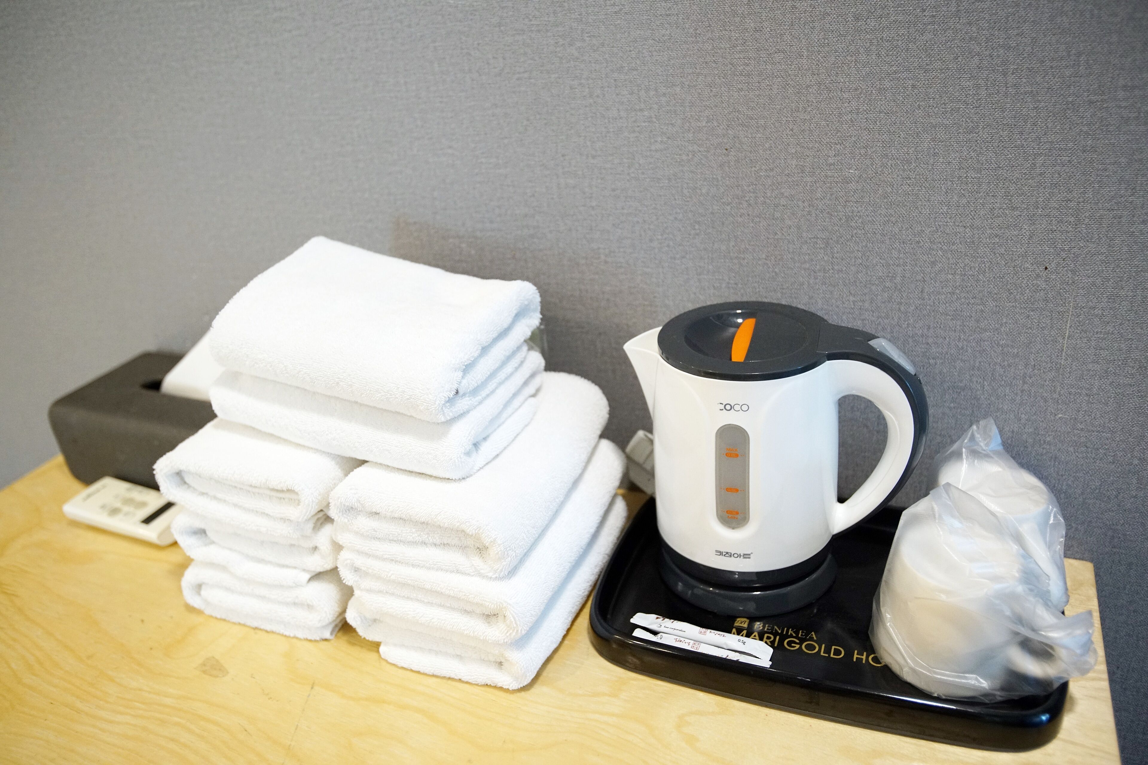A stack of white towels, a white electric kettle, and a plastic bag on a wooden table.