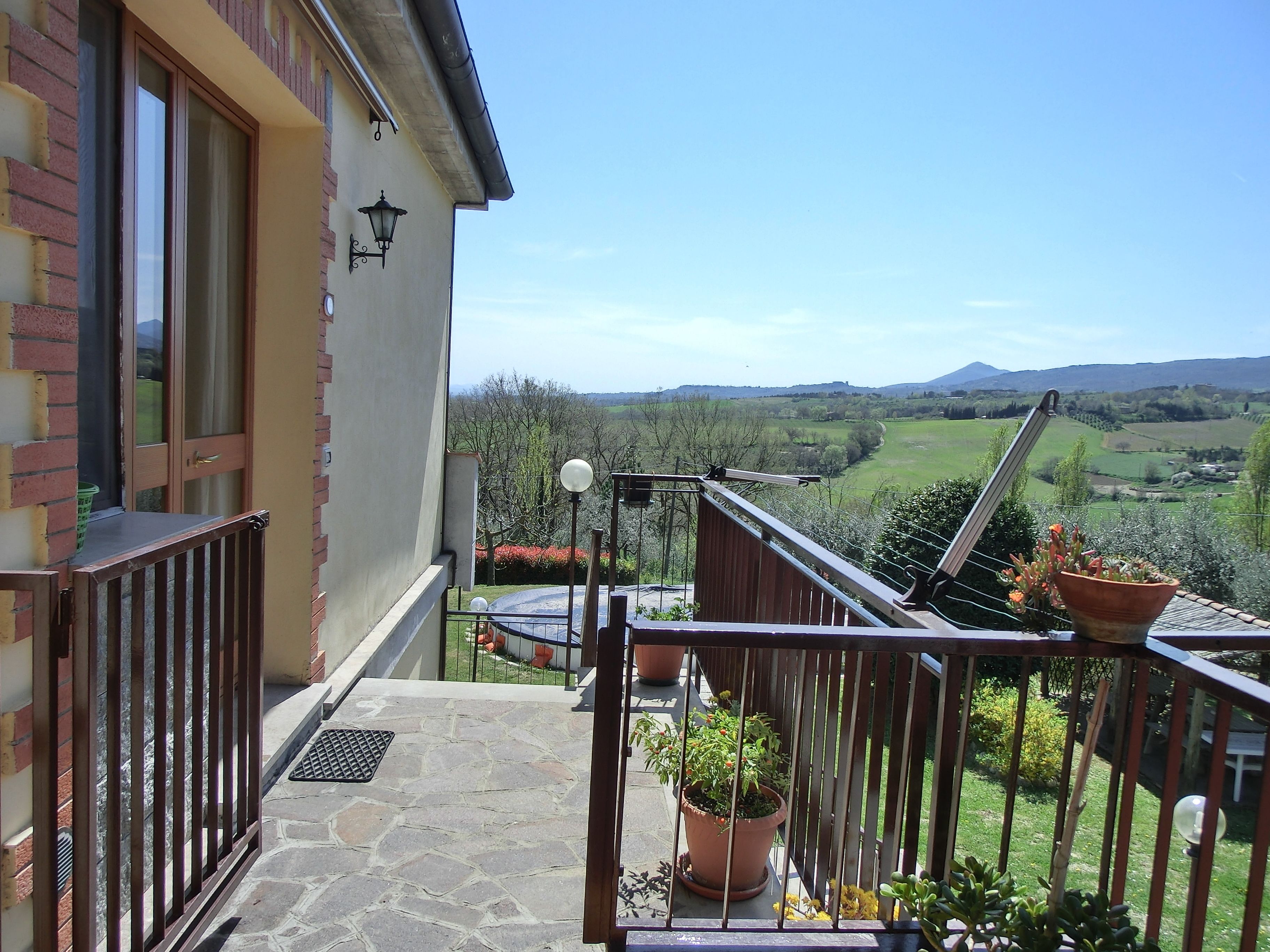A balcony with a view of a green valley and mountains, a potted plant, and a metal railing.