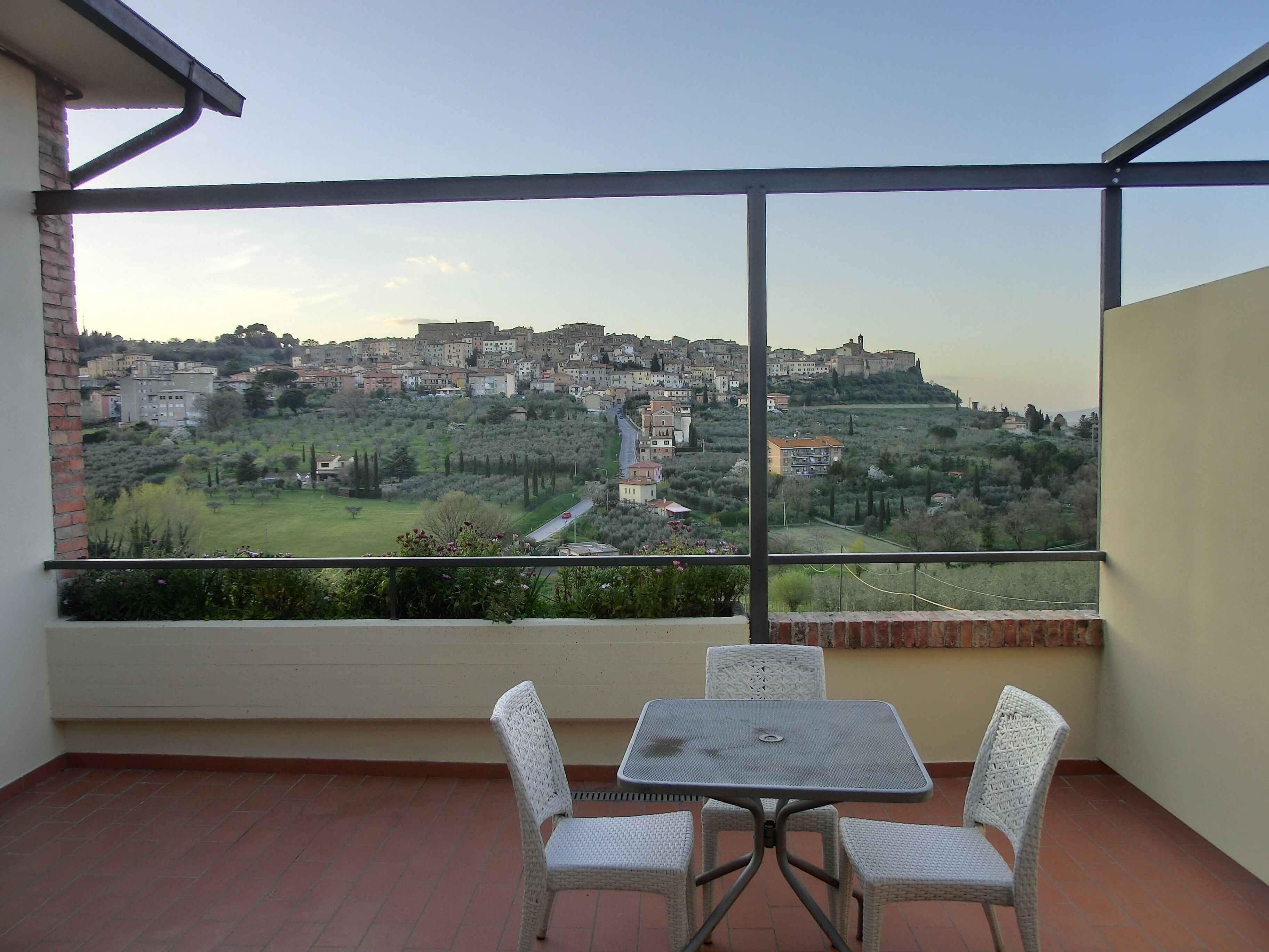 A balcony with a table and chairs, overlooking a town with buildings and greenery.