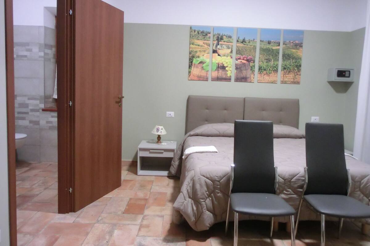 Un dormitorio con cama, dos sillas, una mesita de noche, una lámpara y un aire acondicionado de pared.