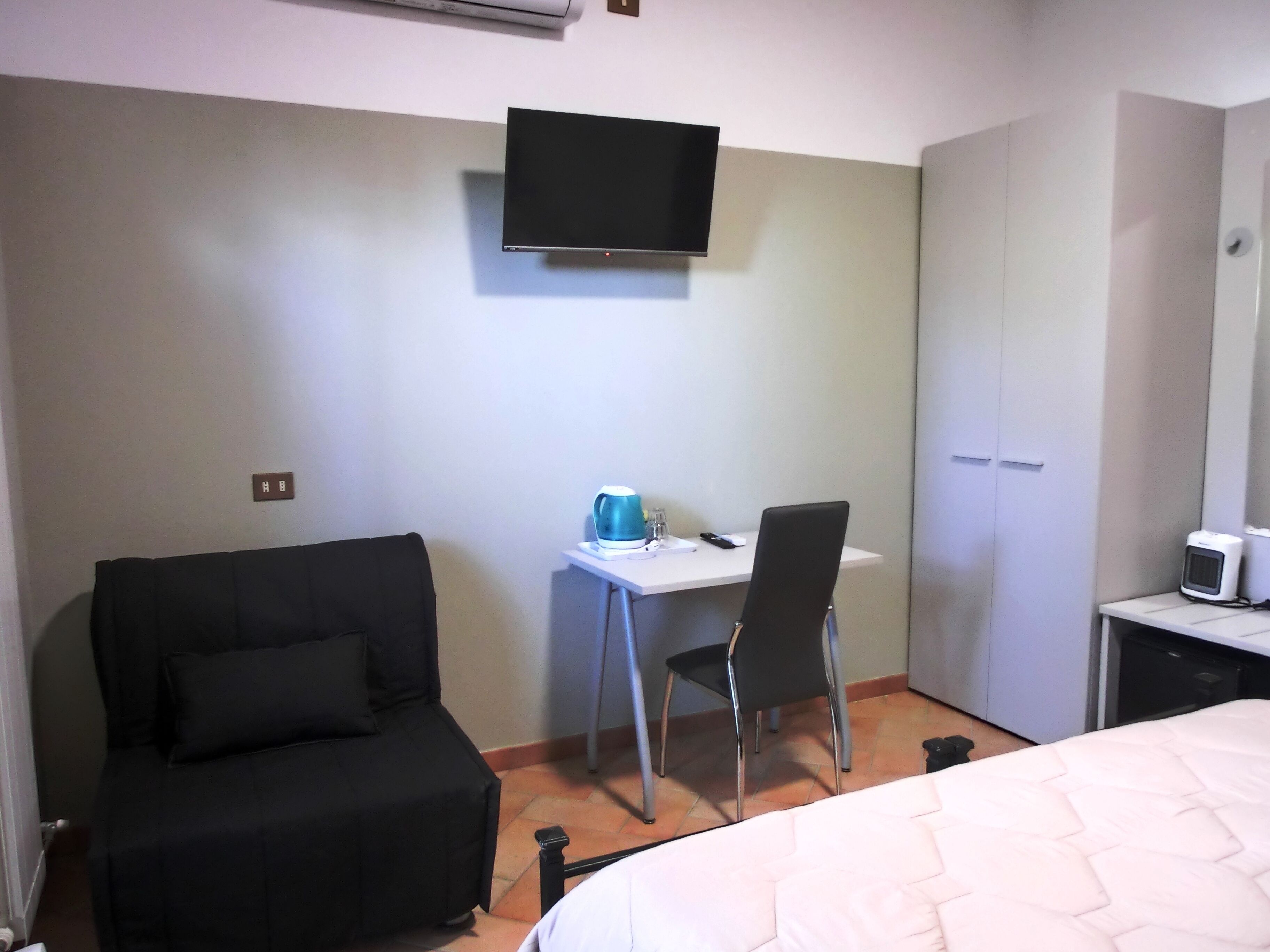 A hotel room with a bed, a desk with a chair, a TV mounted on the wall, and a wardrobe.