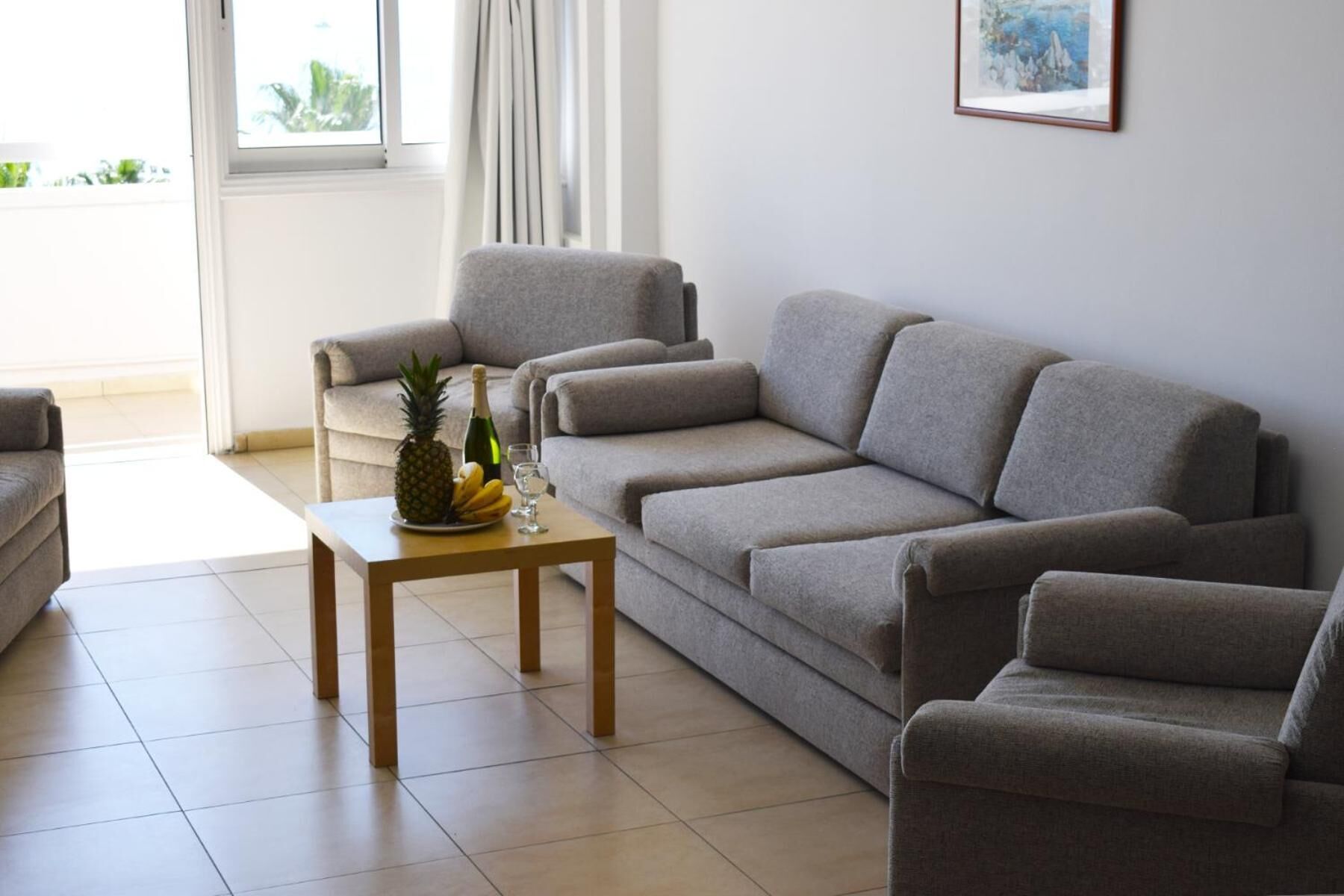 A living room with a grey sofa, a matching armchair, and a wooden coffee table with fruits and a bottle.