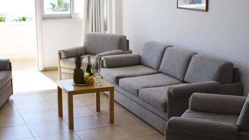 A living room with a grey sofa, a matching armchair, and a wooden coffee table with fruits and a bottle.