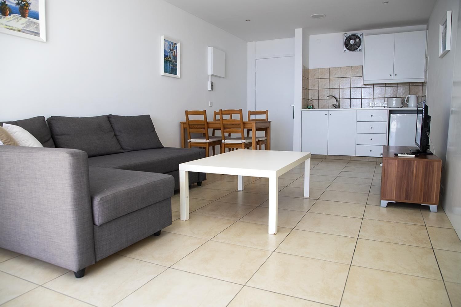 En moderne stue med en grå sofa, et hvidt sofabord og et spiseområde med træstole. Et køkken med hvide skabe og et flisebeklædt bagpanel er synligt i baggrunden.