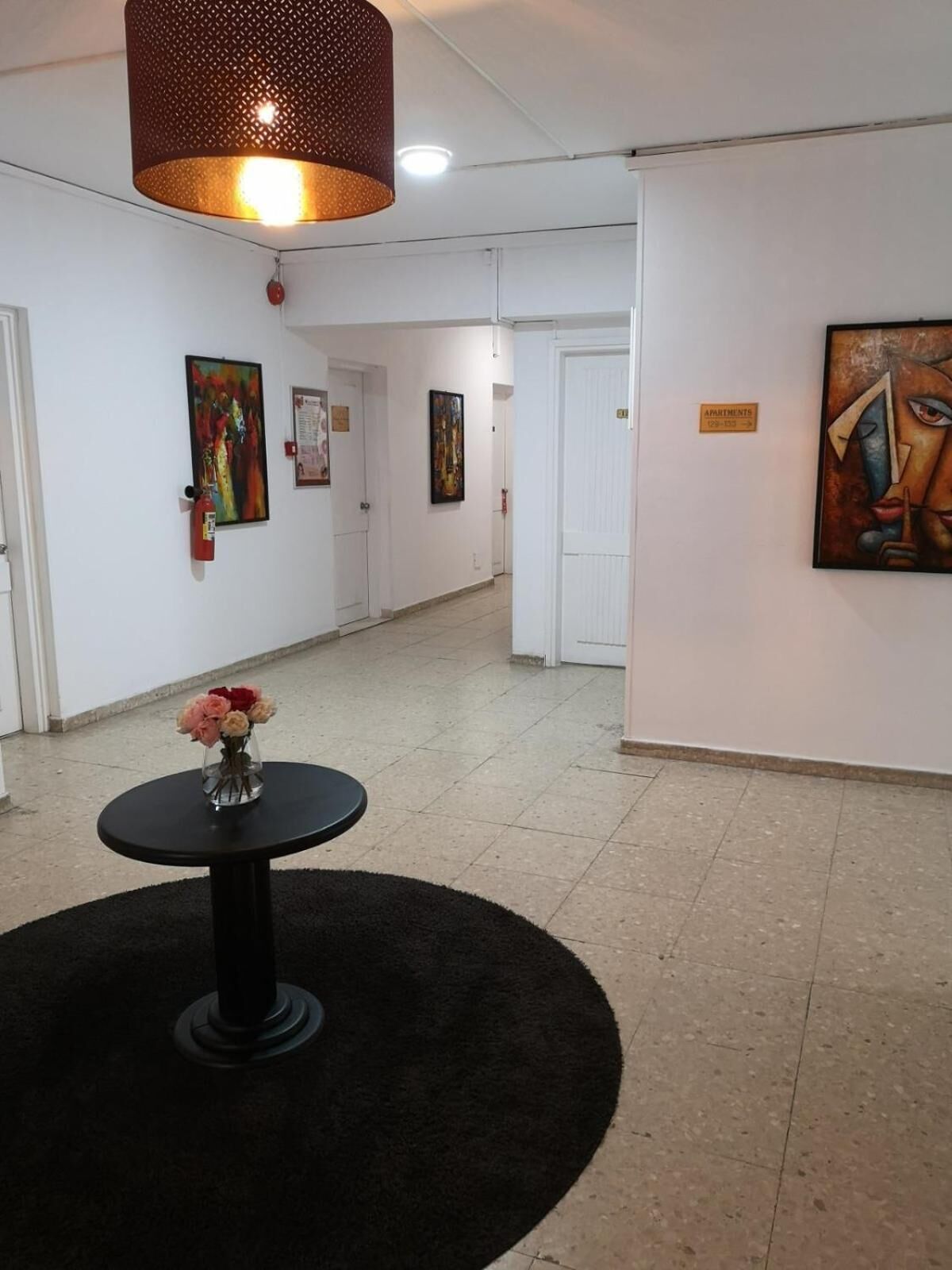 A hallway with white walls, a black circular rug, a round table with a vase of flowers, and several paintings.